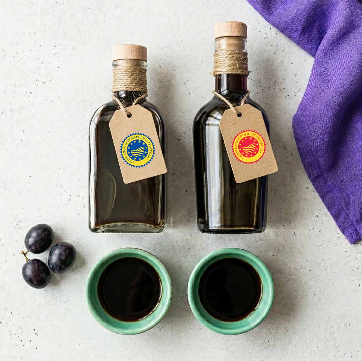 Overhead-Draufsicht auf zwei Balsamico-Flaschen mit angehängten Papierlabels, zwei mintgrünen Schälchen mit Essig, dunklen Weintrauben und einem violetten Leinentuch auf Beton.