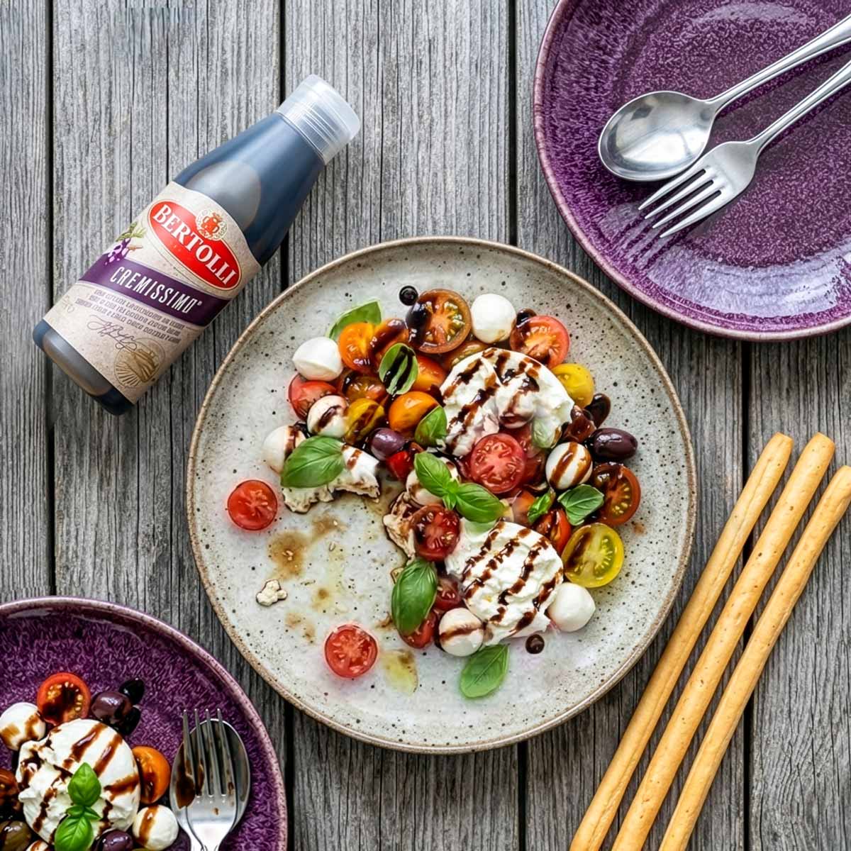 Vogelperspektive einer sommerlichen Mahlzeit: Ein heller Teller mit Tomate-Mozzarella-Salat, Basilikum und Grissini auf einem grauen Holztisch, daneben eine liegende Flasche Bertolli Balsamico Creme und zwei lila Platzteller mit Besteck.