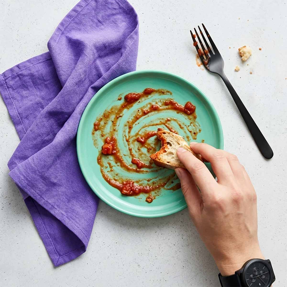 90-Grad-Draufsicht auf eine Hand mit schwarzer Armbanduhr, die ein Stück Brot durch Tomatensaucenreste auf einem mintgrünen Teller zieht, daneben ein violettes Leinentuch.