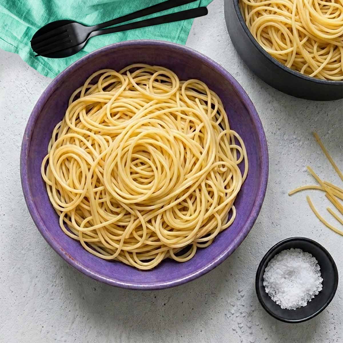 Overhead-Shot einer violetten Schale gef&uuml;llt mit Spaghetti ohne Sauce auf lichtgrauem Beton. Oben rechts ein schwarzer Kochtopf mit Pasta, unten rechts ein schwarzes Sch&auml;lchen mit Salz.