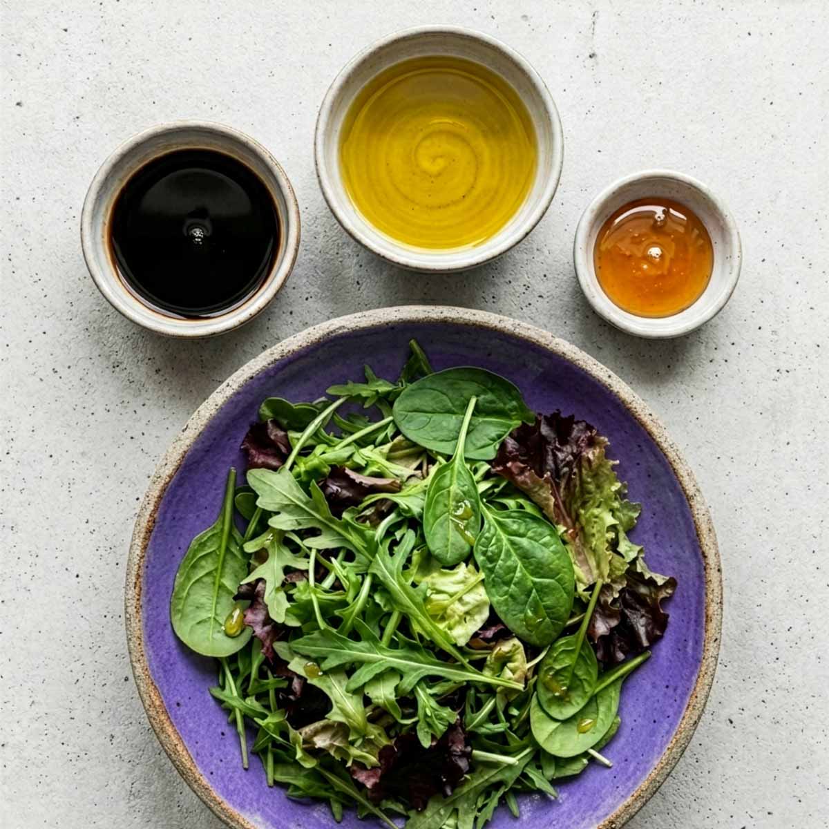 Overhead-Draufsicht auf einen violetten Keramikteller mit Blattsalat, darüber drei Schälchen unterschiedlicher Größe mit Olivenöl, Balsamico-Essig und Honig auf Beton.