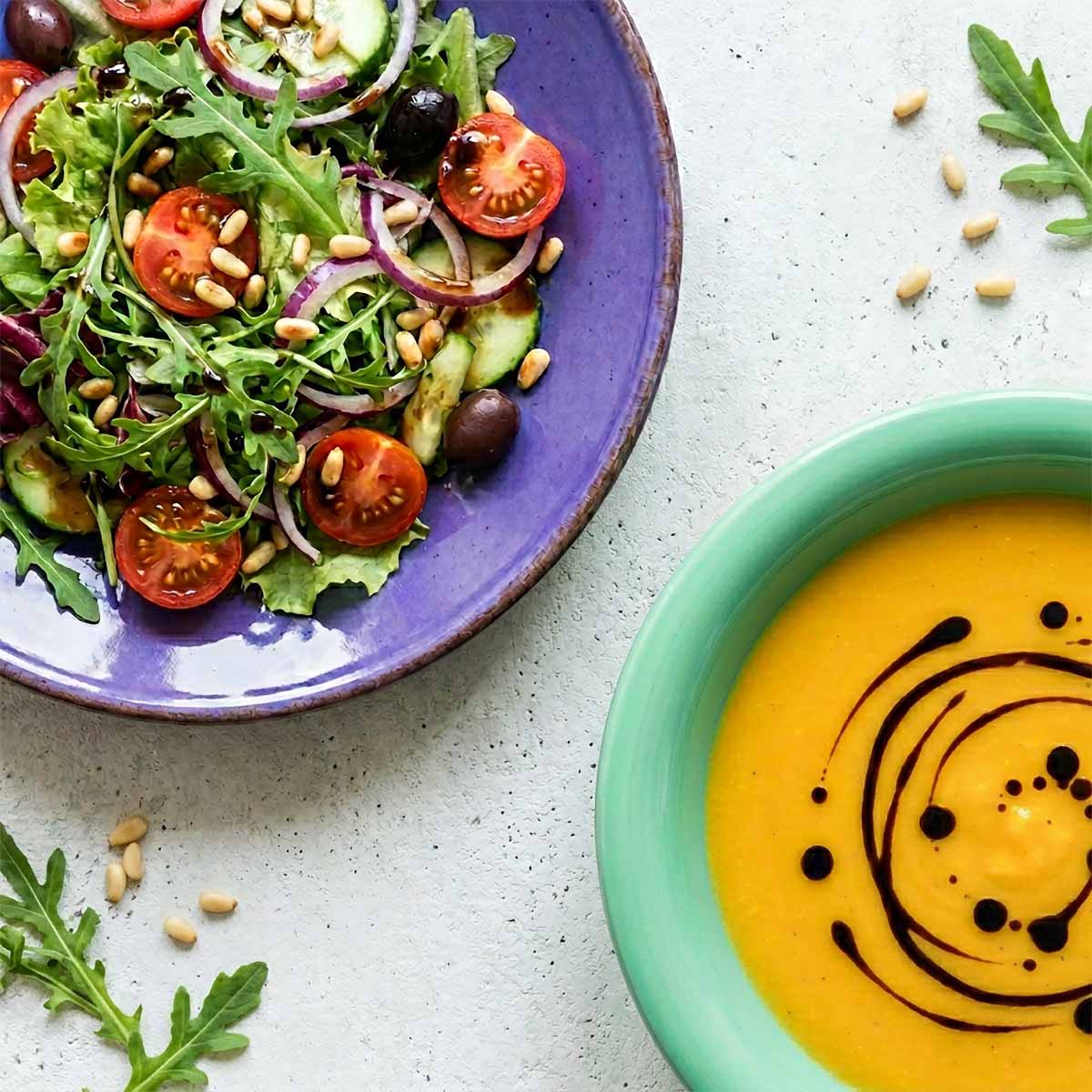 Overhead-Draufsicht auf einen violetten Teller mit gemischtem Salat und einen mintgrünen Teller mit Kürbissuppe, beide mit Balsamico dekoriert, auf hellem Beton.