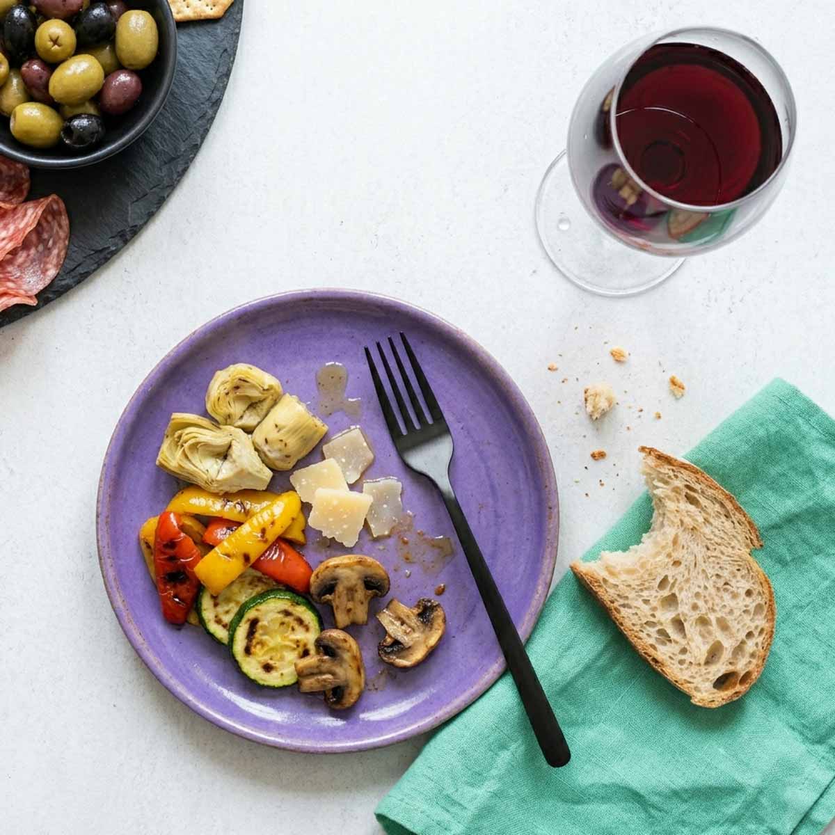 Flache Draufsicht auf einen violetten Keramikteller mit mediterranen Antipasti wie Artischocken, Paprika und Parmesan, daneben ein Rotweinglas, angebissenes Brot und eine Schale Oliven auf hellem Hintergrund.