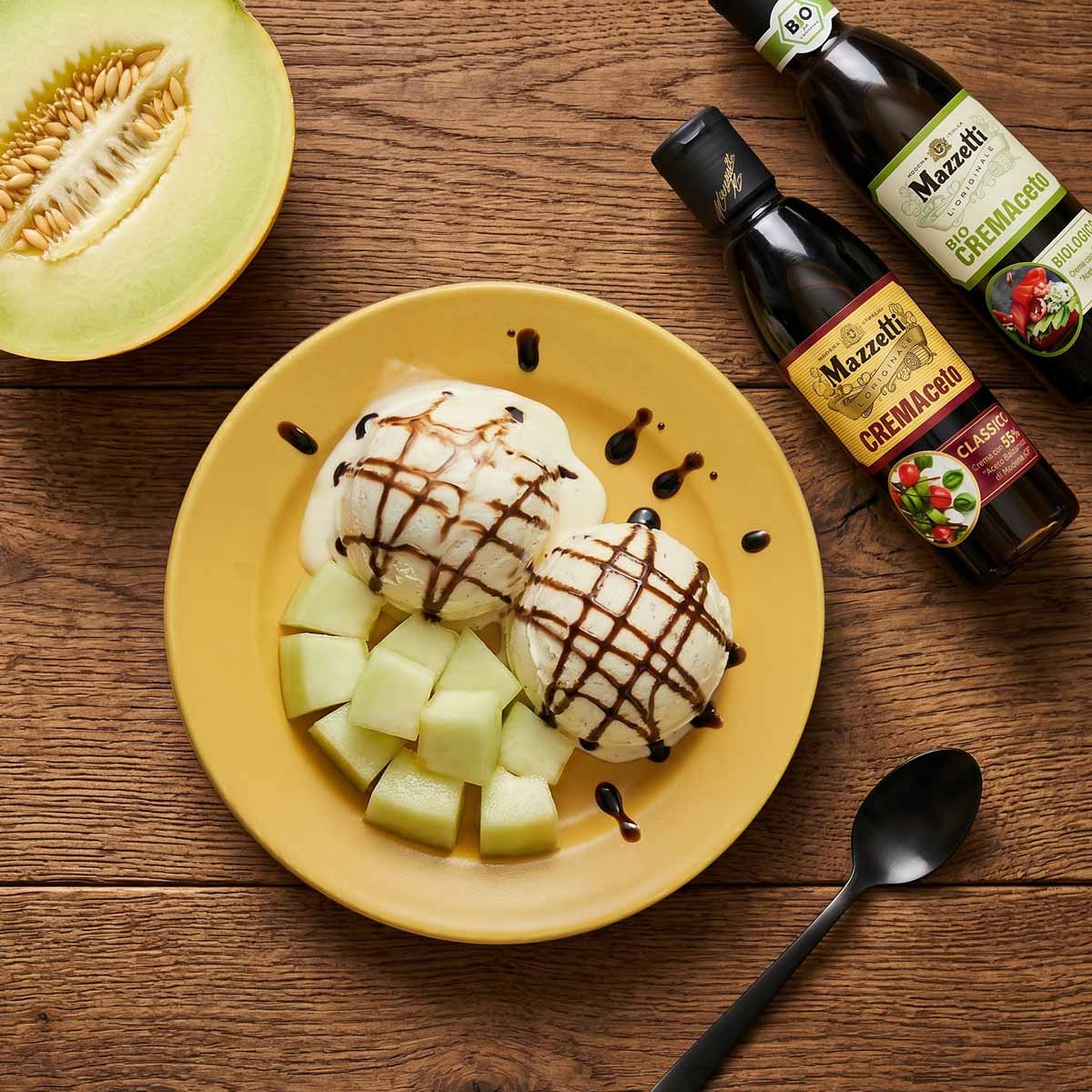 Draufsicht auf einen gelben Teller mit zwei Kugeln Vanilleeis, garniert mit dunkler Mazzetti Cremaceto Balsamico Creme, daneben gewürfelte Honigmelone. Auf einem rustikalen Holztisch liegen zusätzlich eine halbe Honigmelone, ein schwarzer Löffel sowie zwei Flaschen Mazzetti Balsamico Creme (Cremaceto Classico und Biologico).